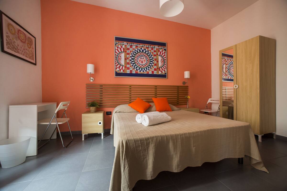 Camera d'albergo moderna con un letto grande, pareti arancioni, una testiera in legno e un elemento decorativo a parete.