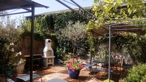 Een terras buiten met houten vloer, een barbecue en diverse plantenpotten.