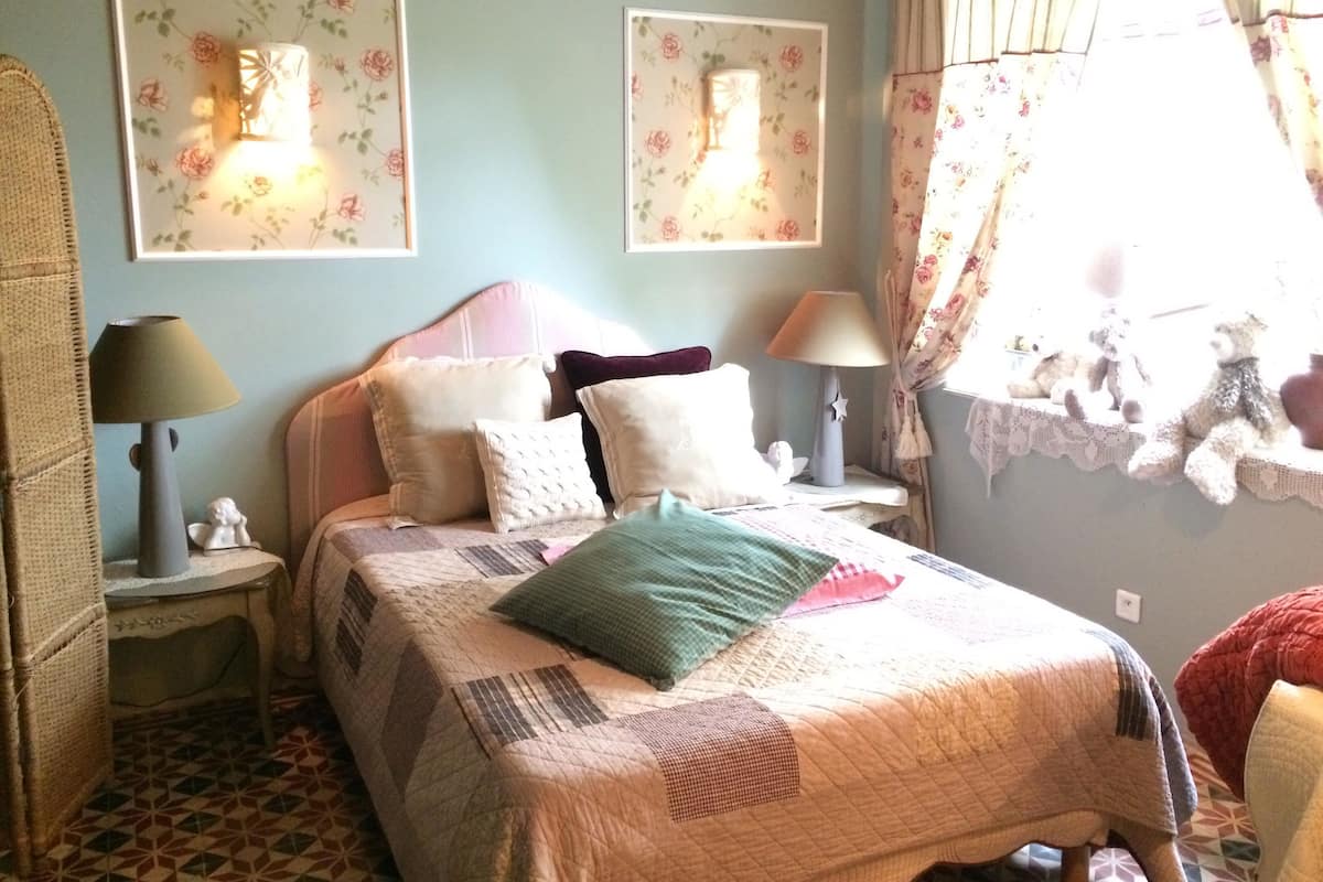 Une chambre à coucher avec un lit, deux lampes de chevet, une table de nuit, une fenêtre avec des rideaux et des tableaux floraux sur les murs.