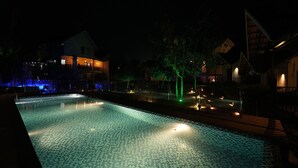 En oplyst swimmingpool med flisebelagte omgivelser og tilstødende bygninger.