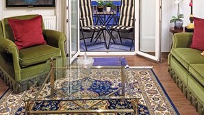 A living room with two green armchairs, a coffee table, and a painting of a ballerina. There is a dining area with striped chairs and a table, and a view of a building with blue awnings. - Tiepolo Galleria Palatina (Thessaloniki)