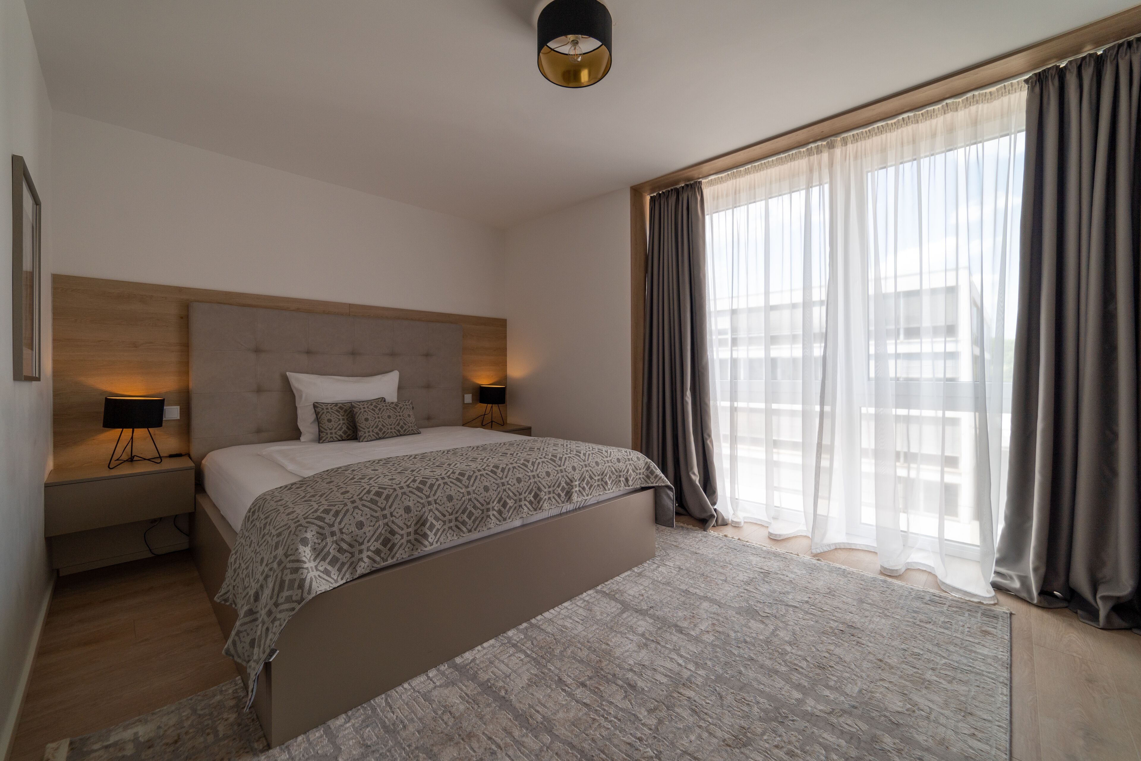 A modern hotel room with a large bed, a wooden headboard, a bedside table with a lamp, a large window with curtains, and a view of the city.