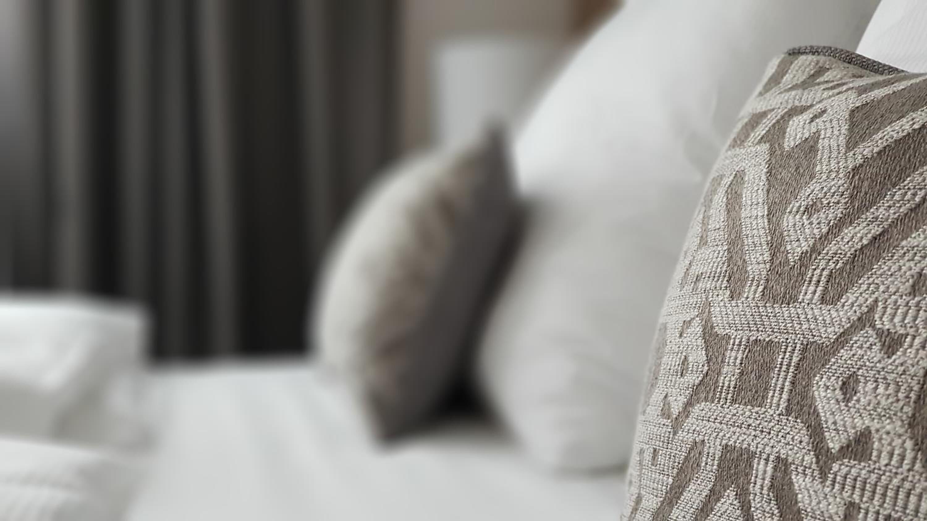 Primer plano de un cojín estampado sobre una cama con cabecera blanca y cortina a rayas al fondo.