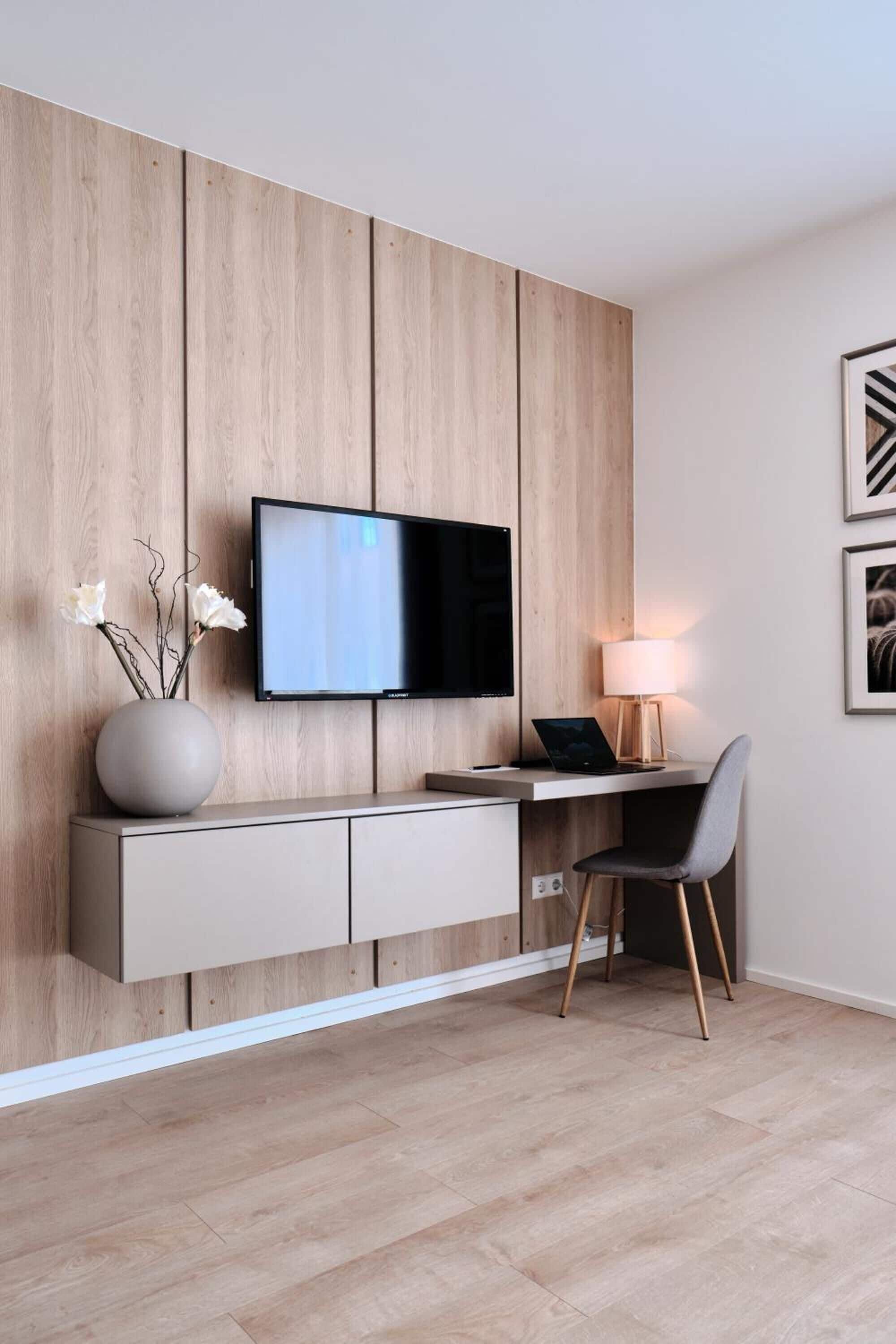 Una sala de estar moderna con un televisor de pantalla plana montado en una pared de madera, un escritorio con una computadora portátil, una silla y un jarrón con flores.