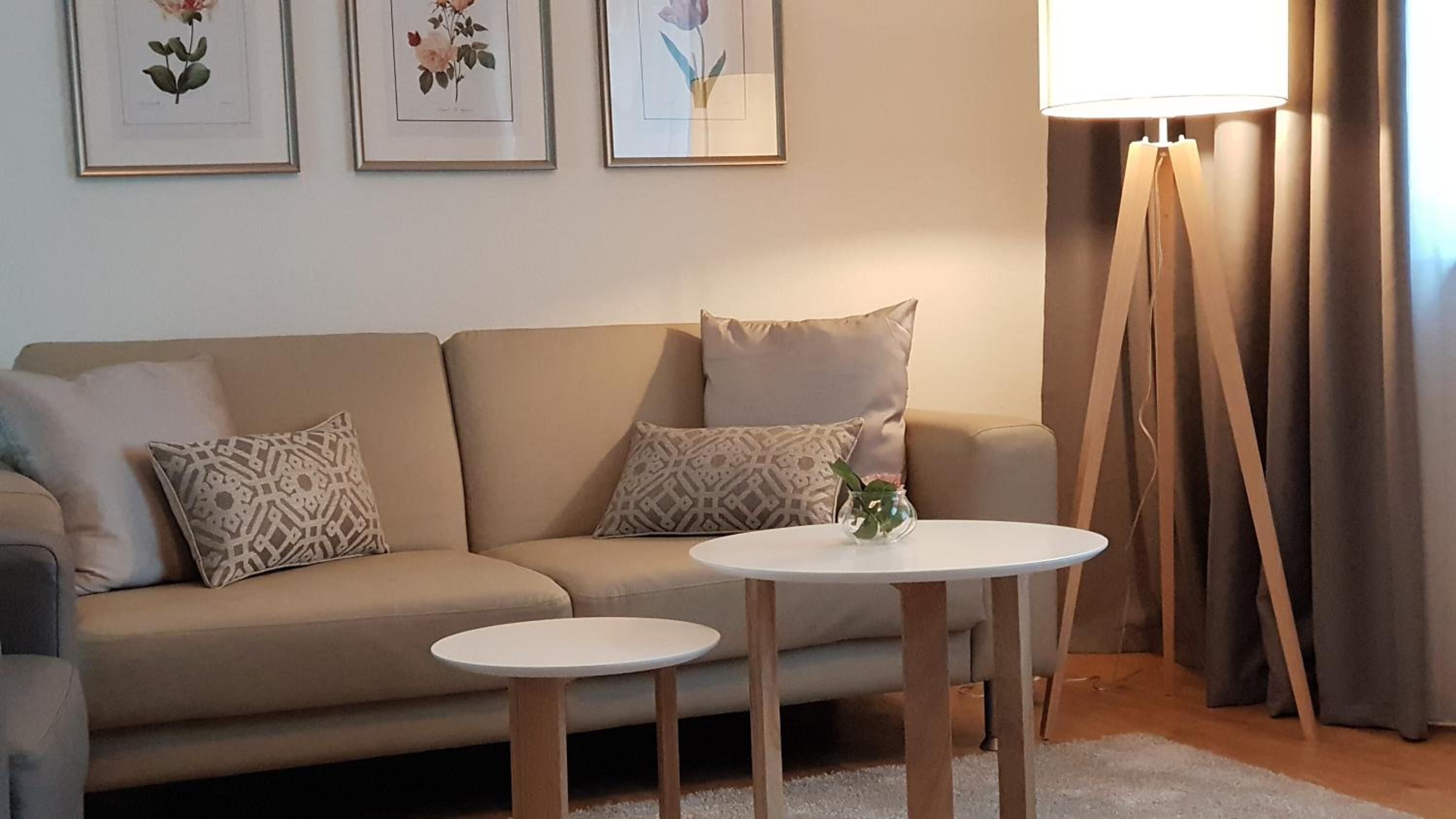 Una sala de estar con un sofá color beige, dos almohadones estampados, una mesita redonda con un florero, y tres láminas botánicas enmarcadas en la pared.