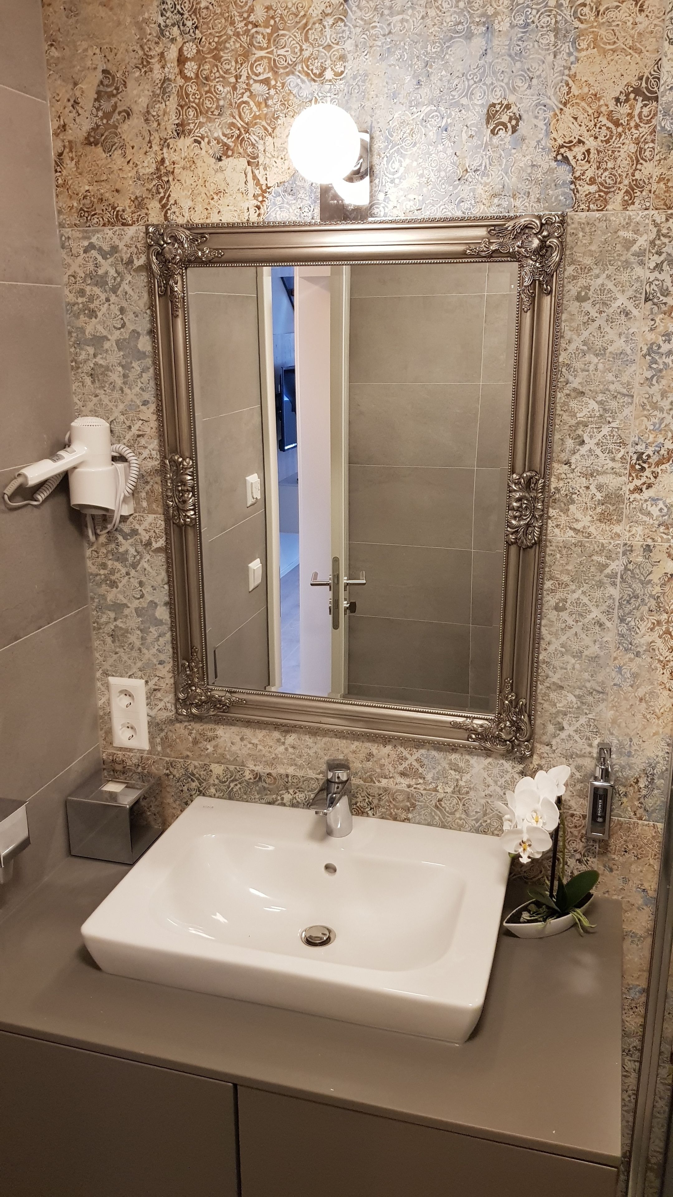 Un baño con un espejo grande y ornamentado, un lavamanos blanco y una orquídea decorativa.