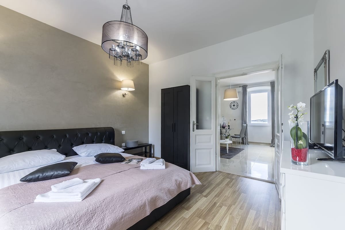 A modern bedroom with a bed, bedside tables, a chandelier, and a view into another room.