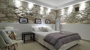 Un dormitorio con pared de piedra, una cama con colcha blanca y morada, una mesita de noche, una silla y cuadros enmarcados.