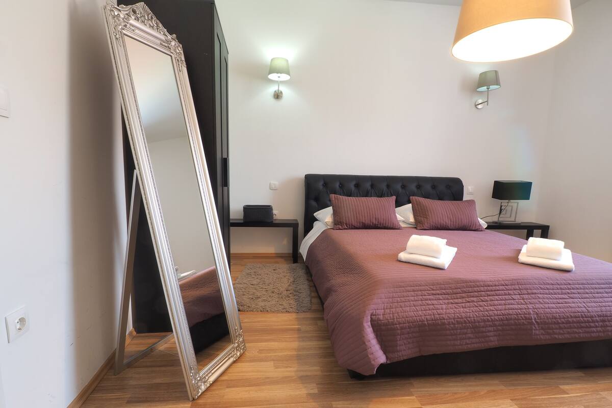 A bedroom with a large mirror, a bed with a headboard, two bedside tables with lamps, and a rug.