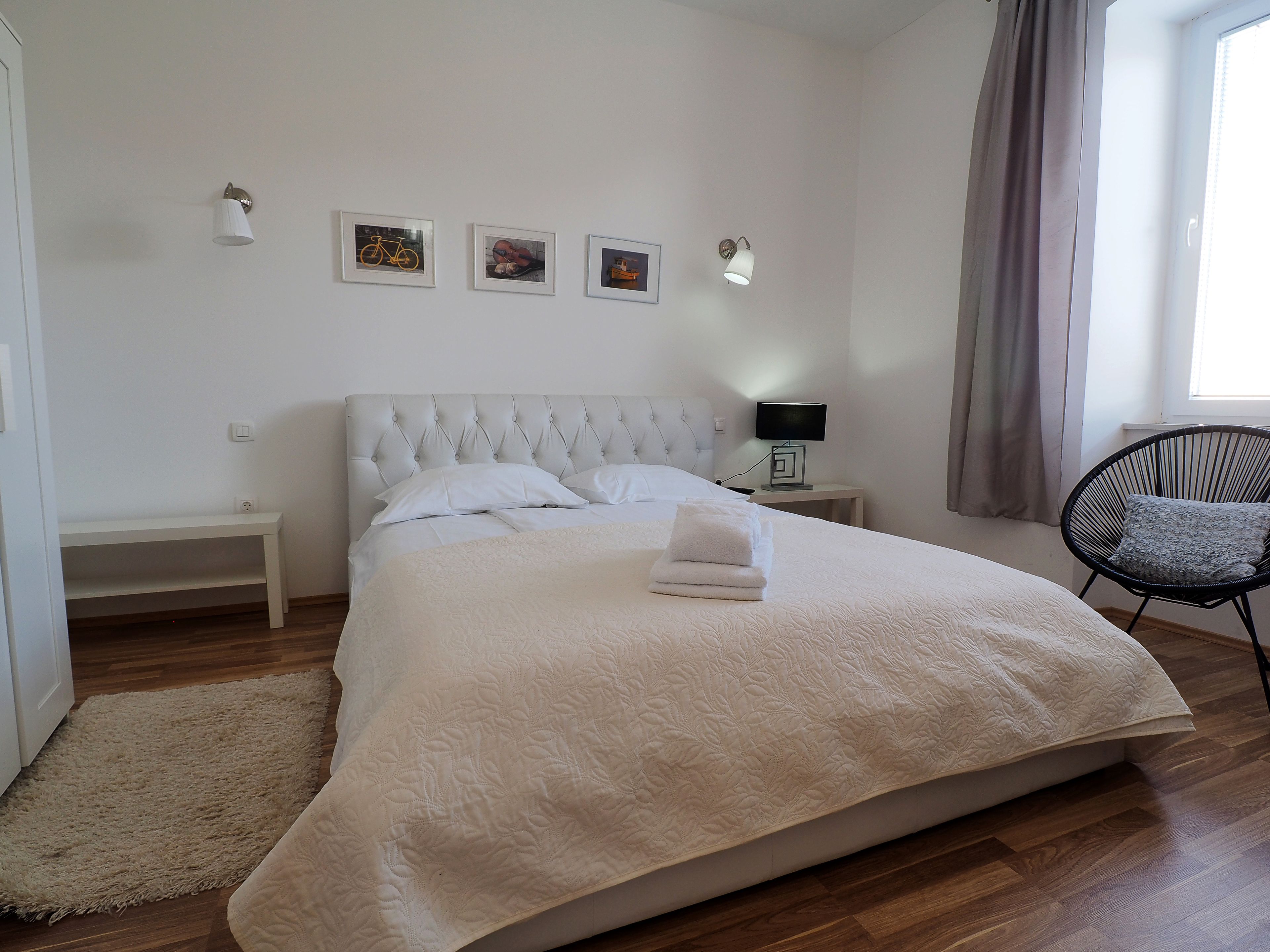 A neatly made bed with a white duvet, a bedside table with a lamp, a chair, and three framed pictures on the wall.
