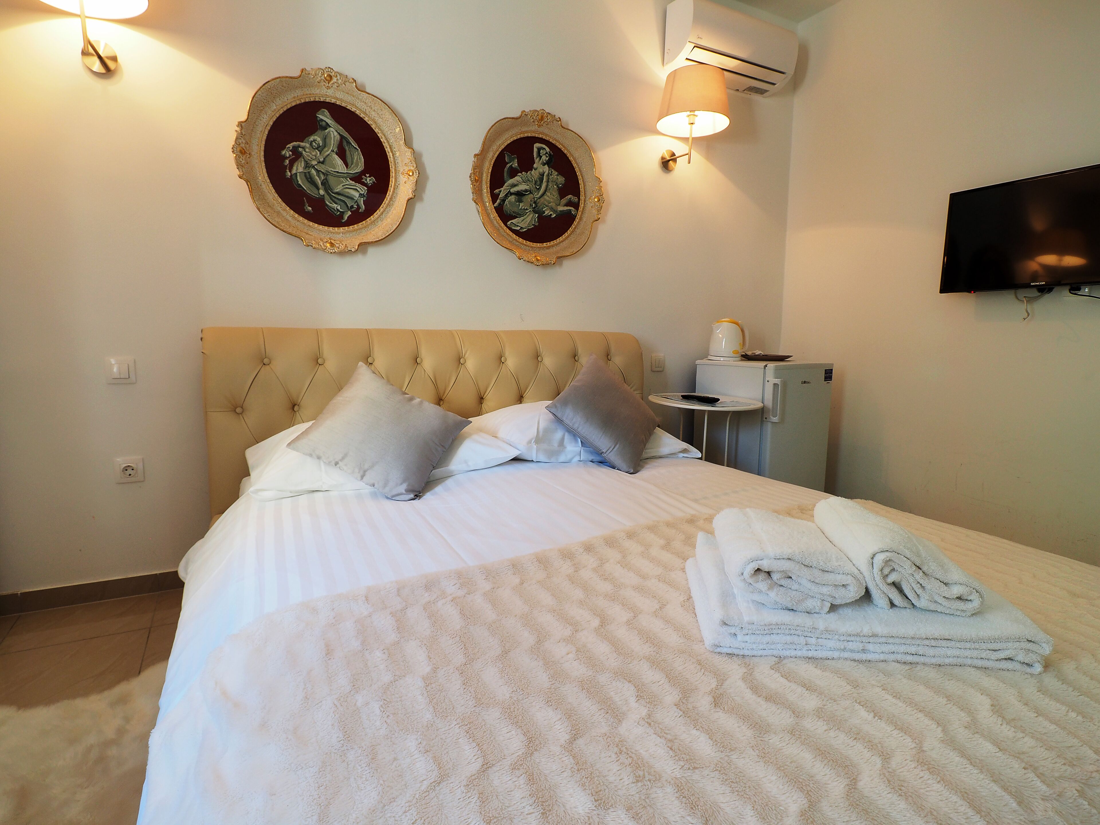A neatly made bed with a tufted headboard, two wall-mounted picture frames, a small table with a lamp, a television, and a refrigerator.