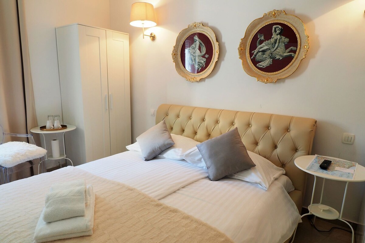 A hotel room with a large bed, two wall-mounted lamps, a wardrobe, a small table with a lamp, and two framed artworks.
