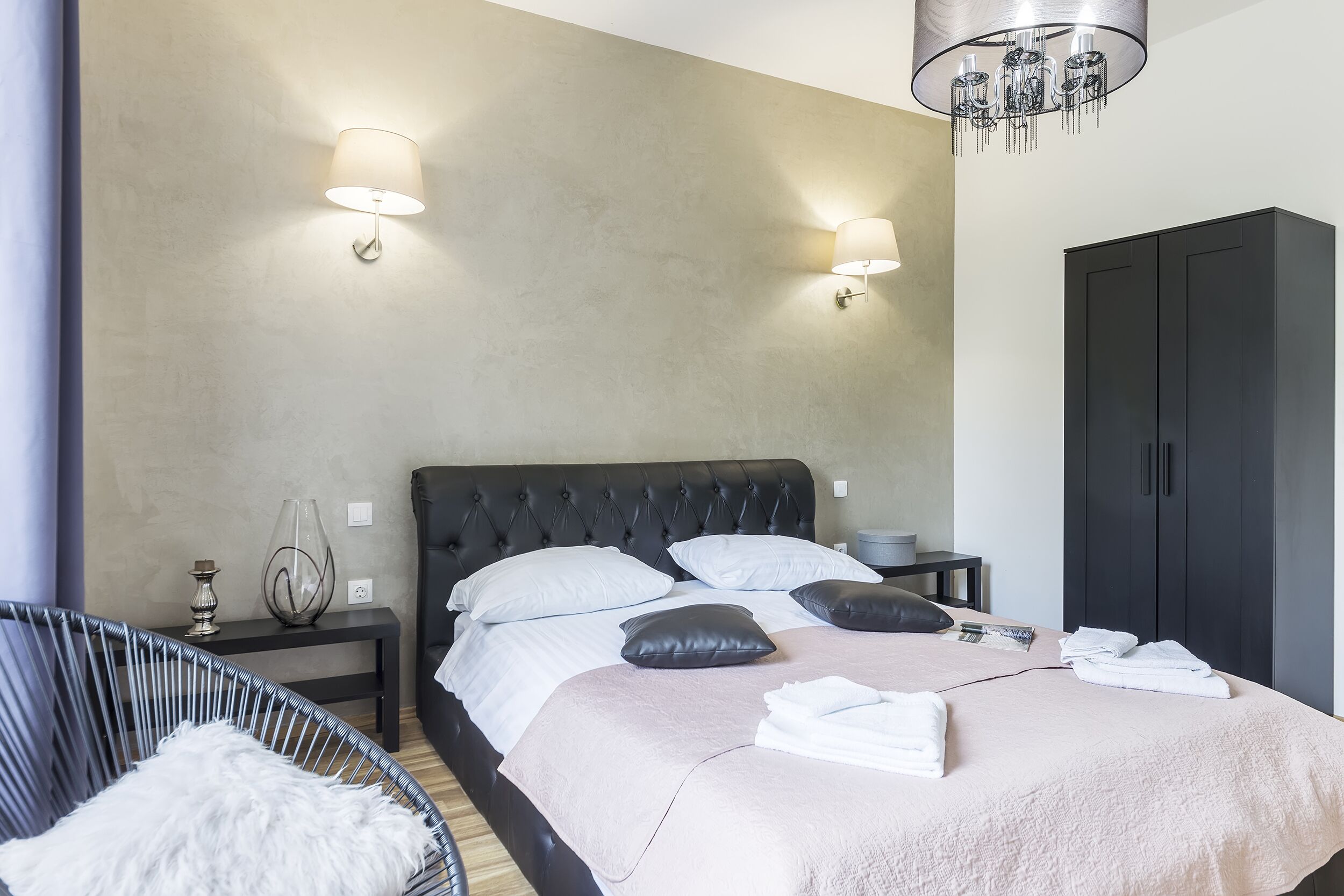 Een net opgemaakt bed met een zwart hoofdbord, twee nachtkastjes met lampen, een donkere garderobe en een rieten stoel met een zacht kussen.