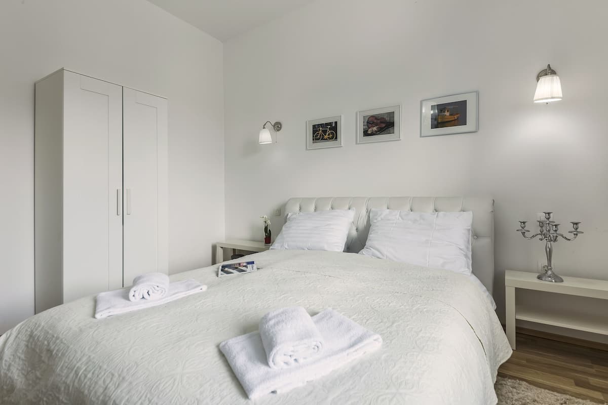 A neatly made bed with white linens, a white wardrobe, and a bedside table with a lamp and a candelabra.