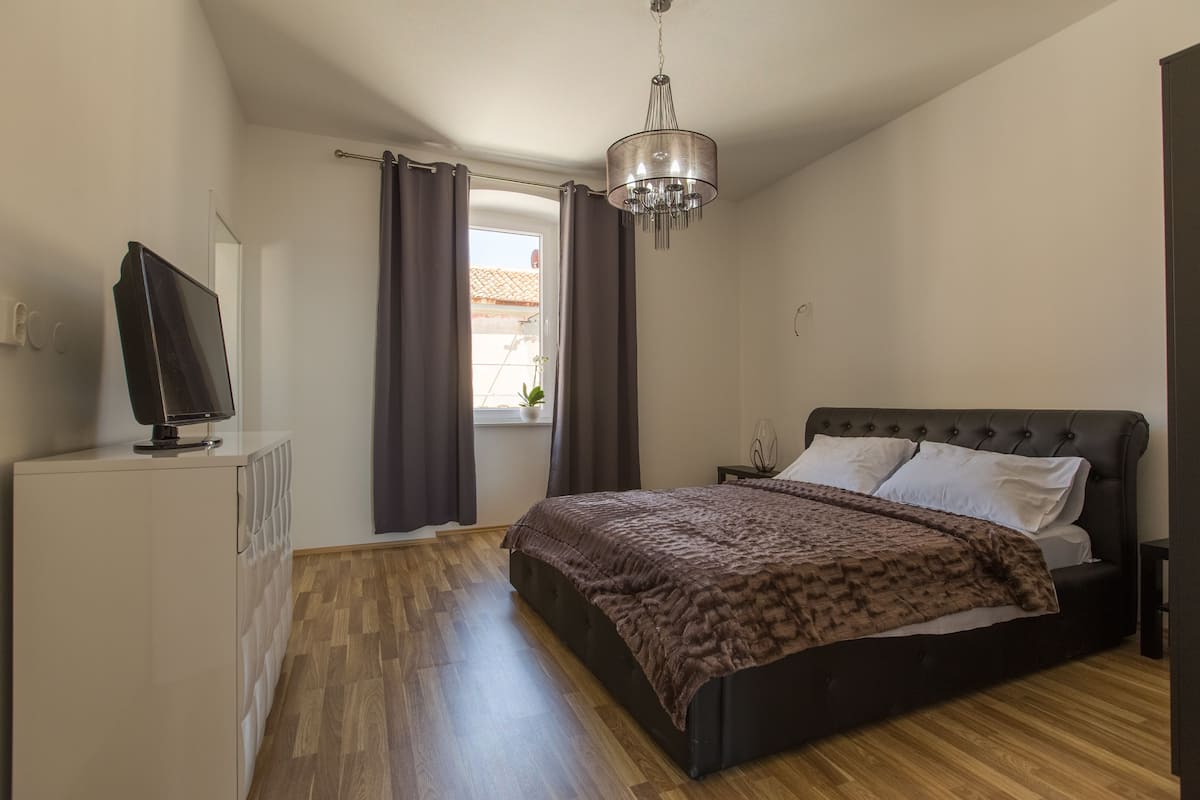 A bedroom with a bed, a television on a cabinet, a chandelier, and a window with curtains.