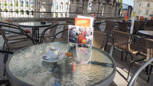 Un café al aire libre con mesas redondas y sillas de mimbre, que incluye una mesa de vidrio con una taza, un platillo y un menú.