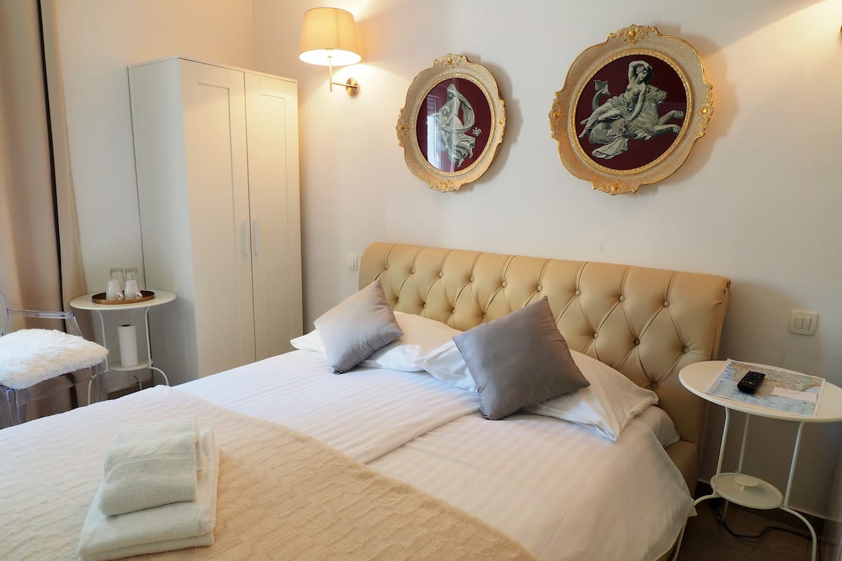 A hotel room with a large bed, two wall-mounted lamps, a small table with a remote control, and a wardrobe.