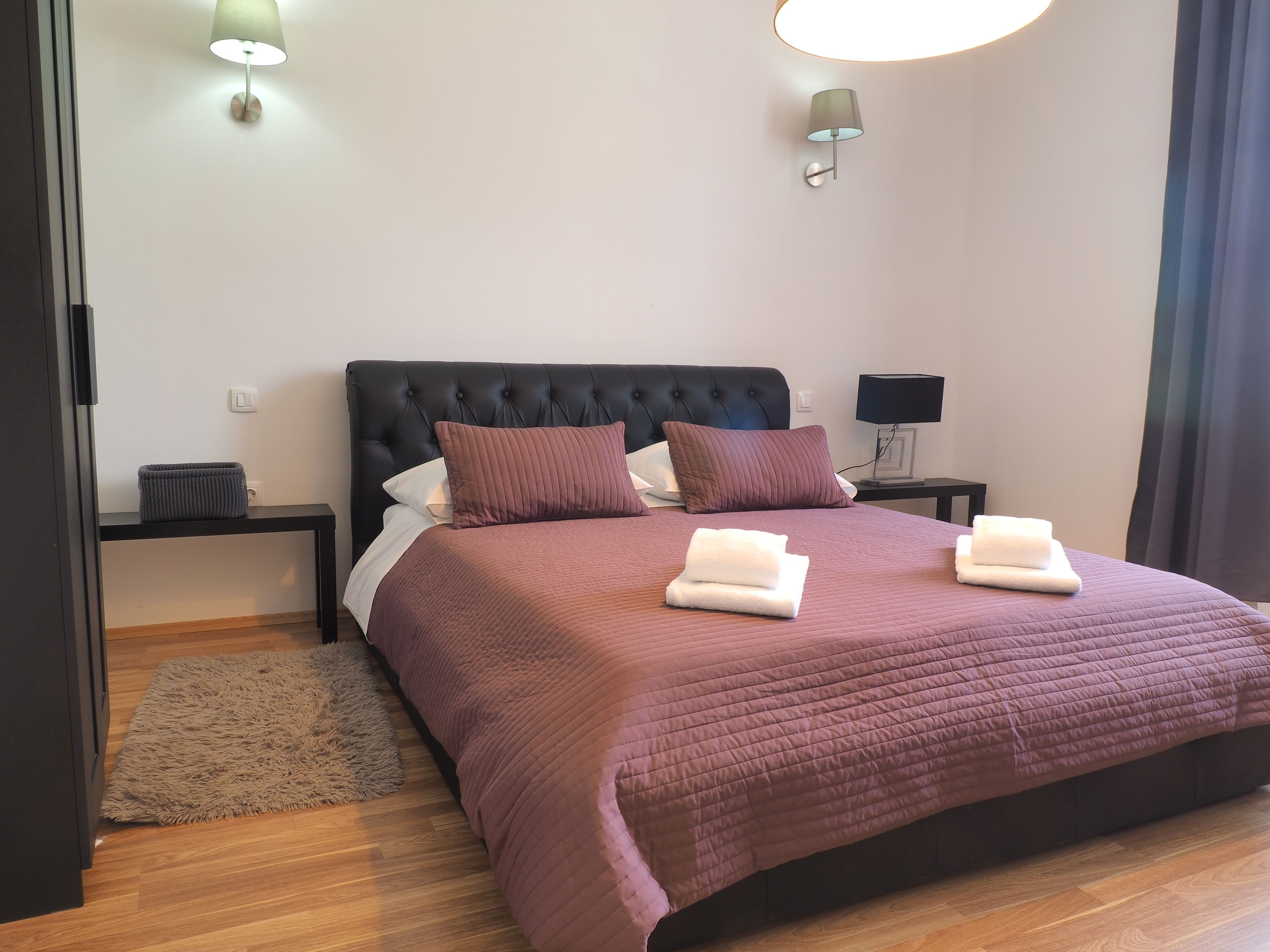 Un lit bien fait, avec une tête de lit sombre, deux tables de chevet avec des lampes, un tapis moelleux et un luminaire fixé au mur.
