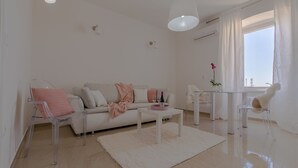 Una sala de estar moderna con un sofá blanco, sillas transparentes, una mesa de centro de vidrio y un ventanal con cortinas.