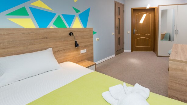 A hotel room with a bed, a wooden headboard, a green bedspread, a white pillow, a white towel, a nightstand, a wall with geometric patterns, a door, a mirror, and a wardrobe. - Blum Hotel (Lviv)