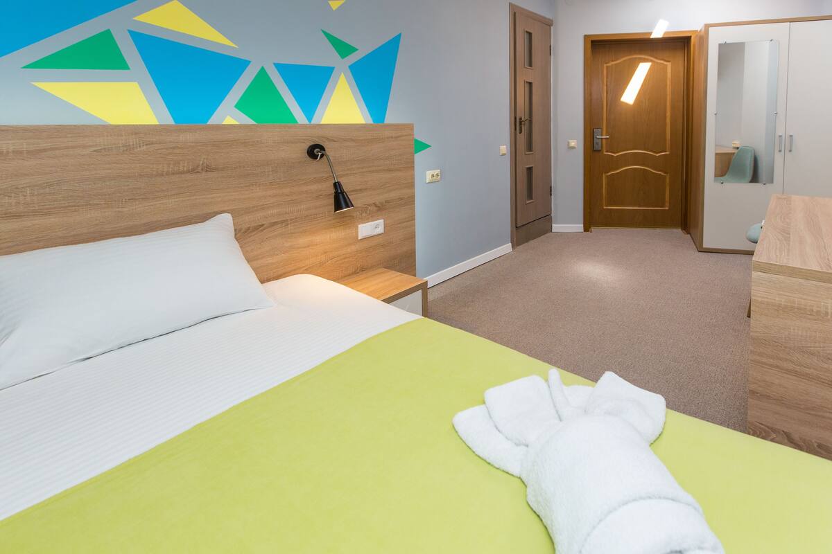 Une chambre d’hôtel avec un lit, une tête de lit en bois, une couvre-lit verte, un oreiller blanc, une serviette blanche, une table de chevet, un mur orné de motifs géométriques, une porte, un miroir et une armoire.