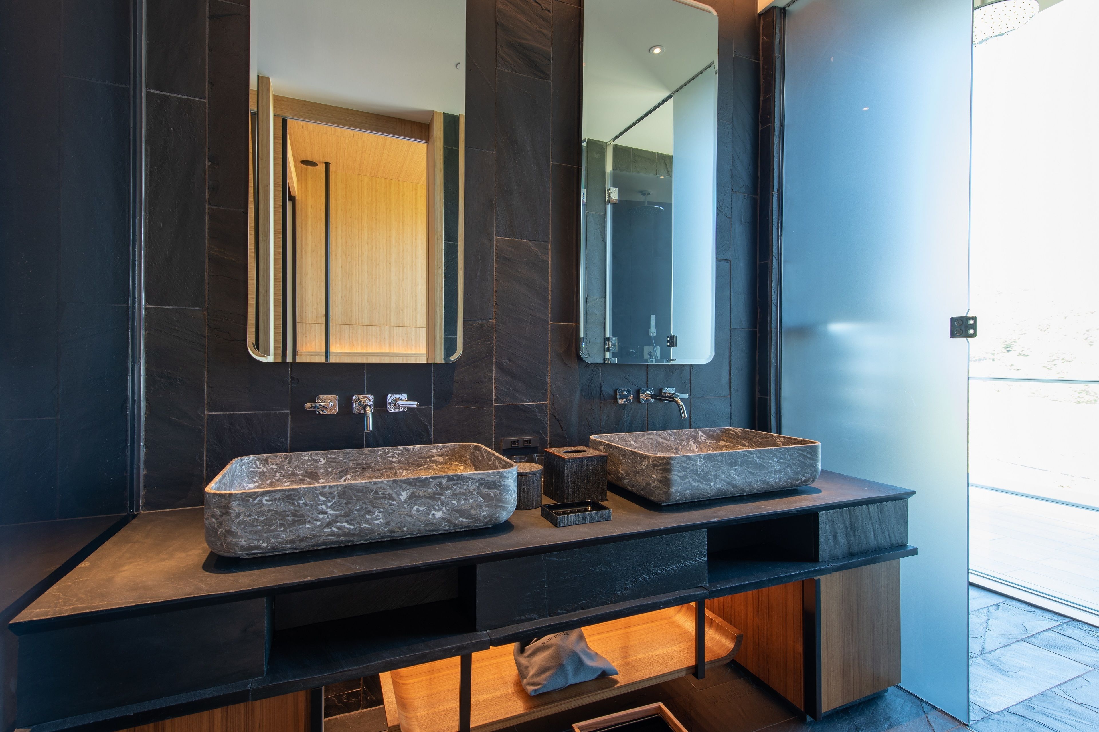 A modern bathroom with two large, rectangular stone sinks, a large mirror, and a wooden vanity.