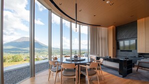 A modern interior with a round dining table, chairs, a sofa, and a TV. A large window offers a view of a mountain. - Skye Niseko (Kutchan)