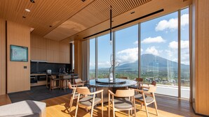 A modern kitchen with wooden cabinetry and a large island, a dining area with wooden chairs and a round table, and large floor-to-ceiling windows offering a view of a mountain and cityscape. - Skye Niseko (Kutchan)