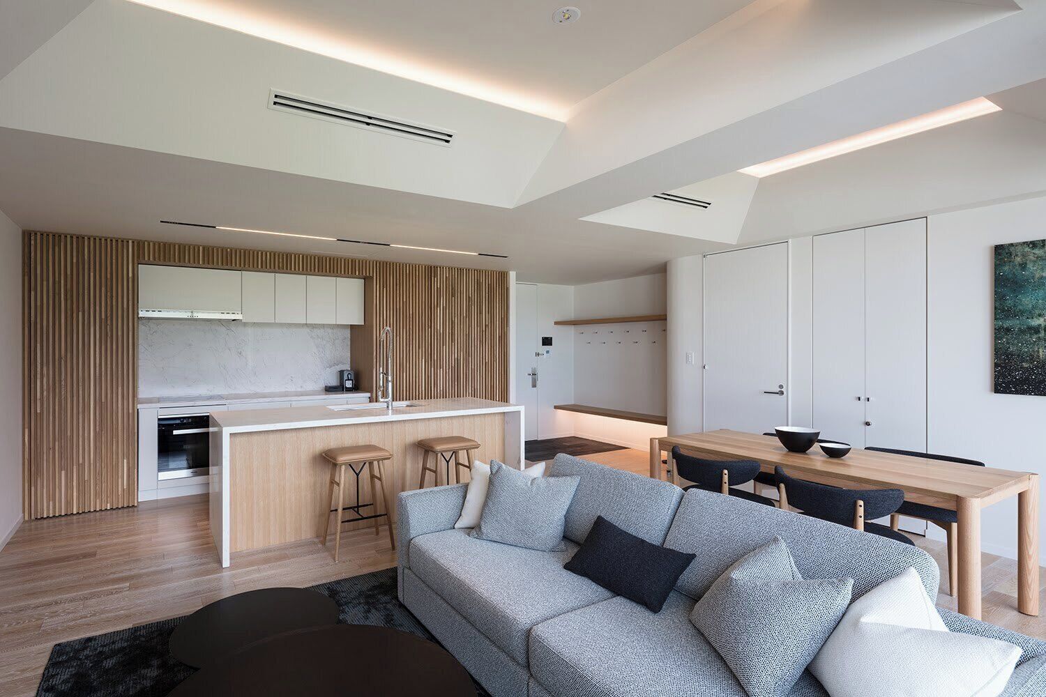 A modern kitchen with a central island, a dining area with a wooden table, and a grey sofa with cushions.