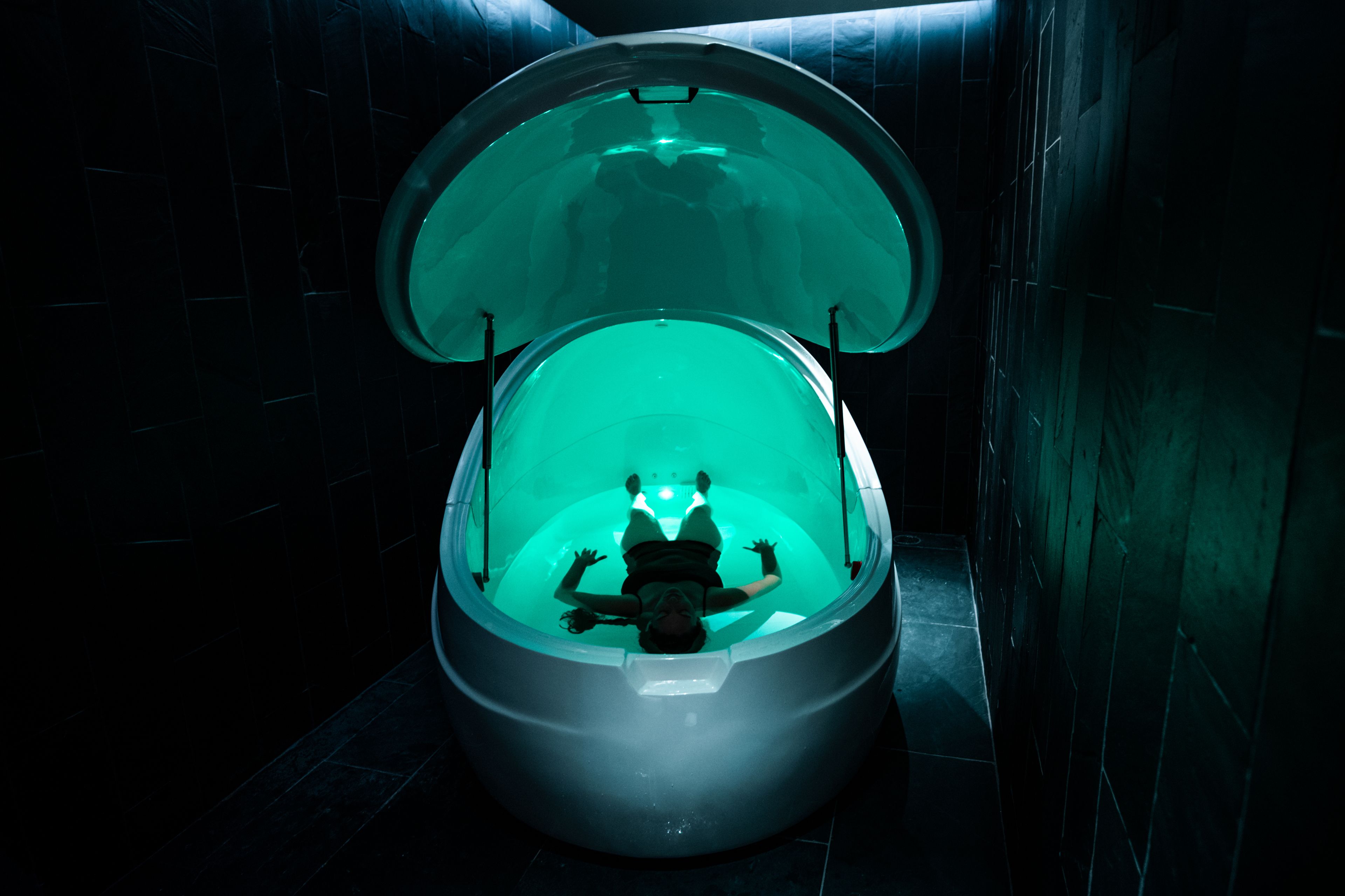 Una persona recostada dentro de una bañera transparente con forma de cúpula y detalles en azul claro, ubicada en una habitación oscura con paredes de azulejos.