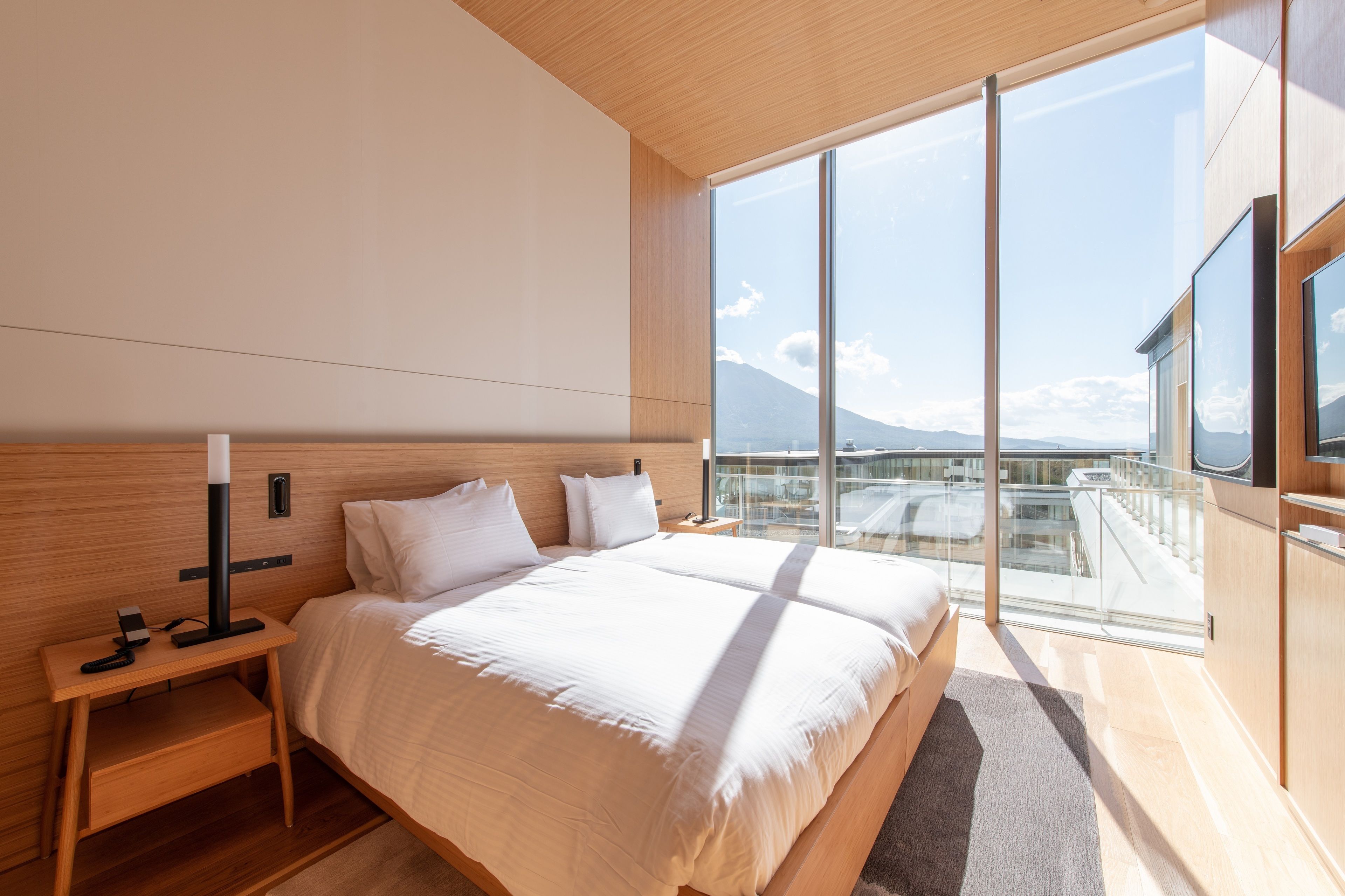 Un dormitorio moderno con una cama grande, una mesita de noche y una mesilla auxiliar. Una ventana grande ofrece vistas a la ciudad y a las montañas.