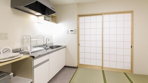Una cocina con un lavamanos, estufa y un reloj. Hay una puerta corredera con diseño de cuadrícula y un tatami en el suelo.