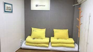 Una habitación pequeña con dos camas, cada una con un almohadón amarillo y una colcha blanca. Hay un cuadro enmarcado en la pared y un equipo de aire acondicionado sobre las camas.