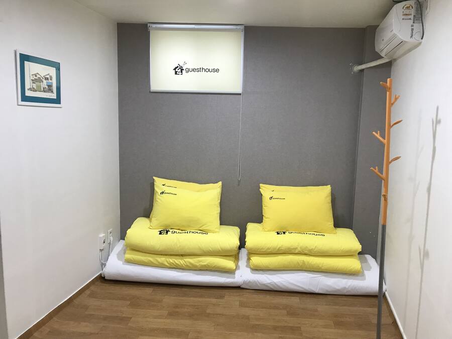 Una habitación pequeña con dos camas, cada una con un almohadón amarillo y una colcha blanca. Hay un cuadro enmarcado en la pared y un equipo de aire acondicionado sobre las camas.