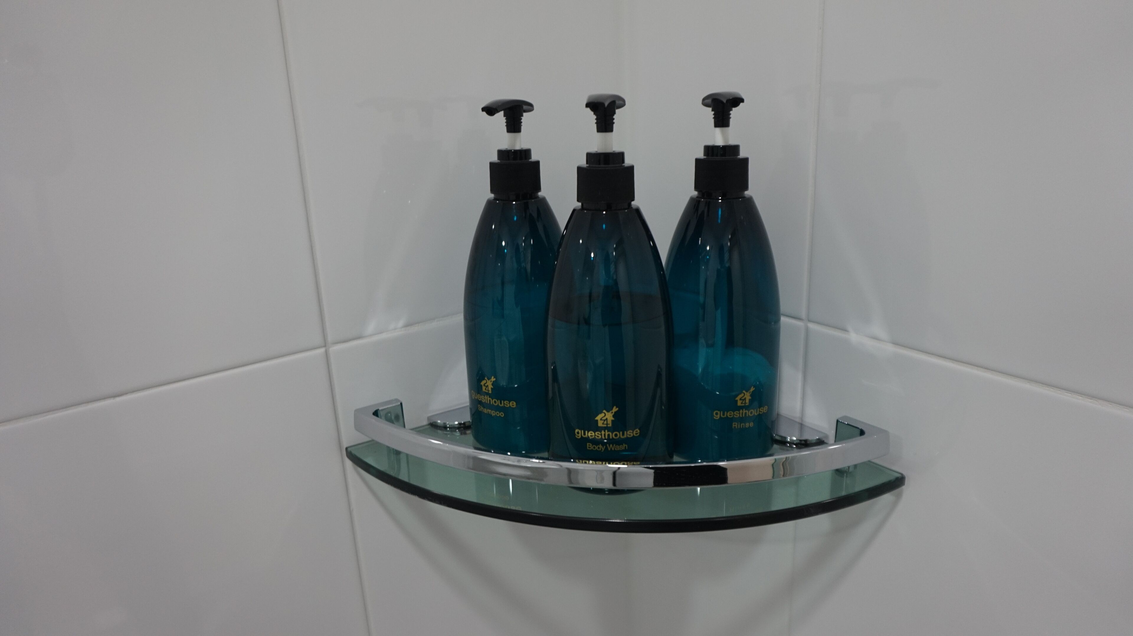 Une salle de bain avec une étagère en verre sur laquelle sont posées trois bouteilles de gel douche et de shampoing, typiques de celles que l’on trouve dans les chambres d’hôtes.