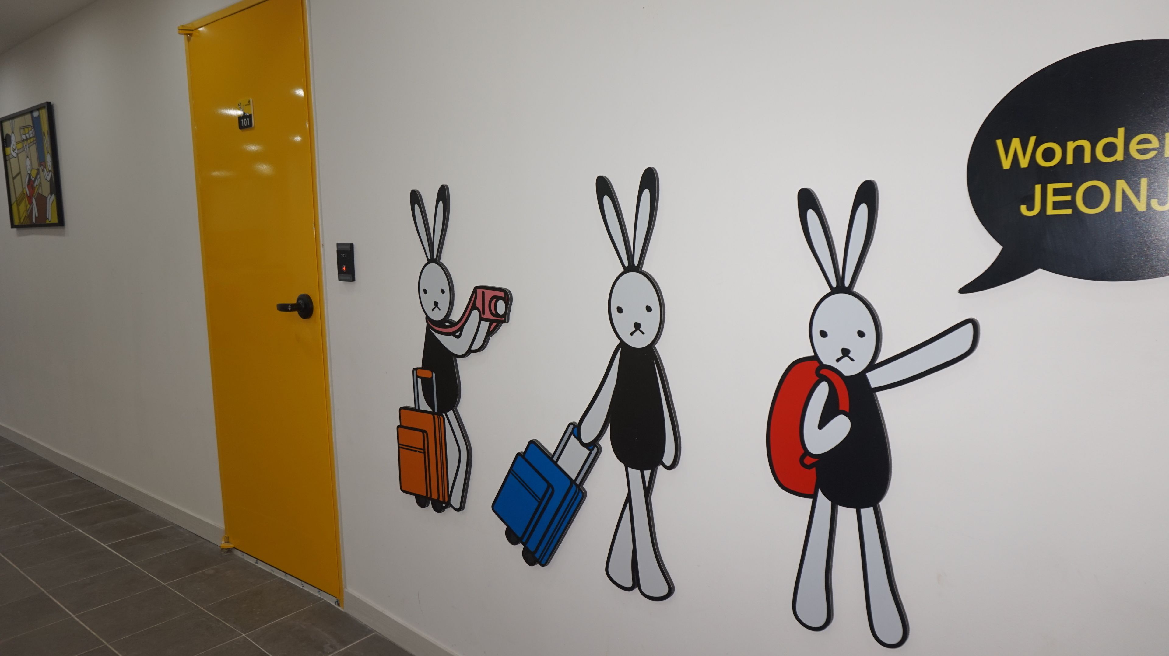 Een gang met een gele deur en een muurschildering van drie cartoon konijnen met koffers.