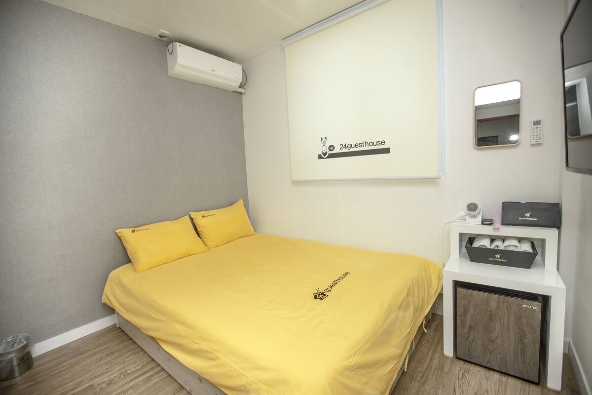A hotel room with a bed, a small white cabinet with a mirror, and a wall-mounted air conditioner.