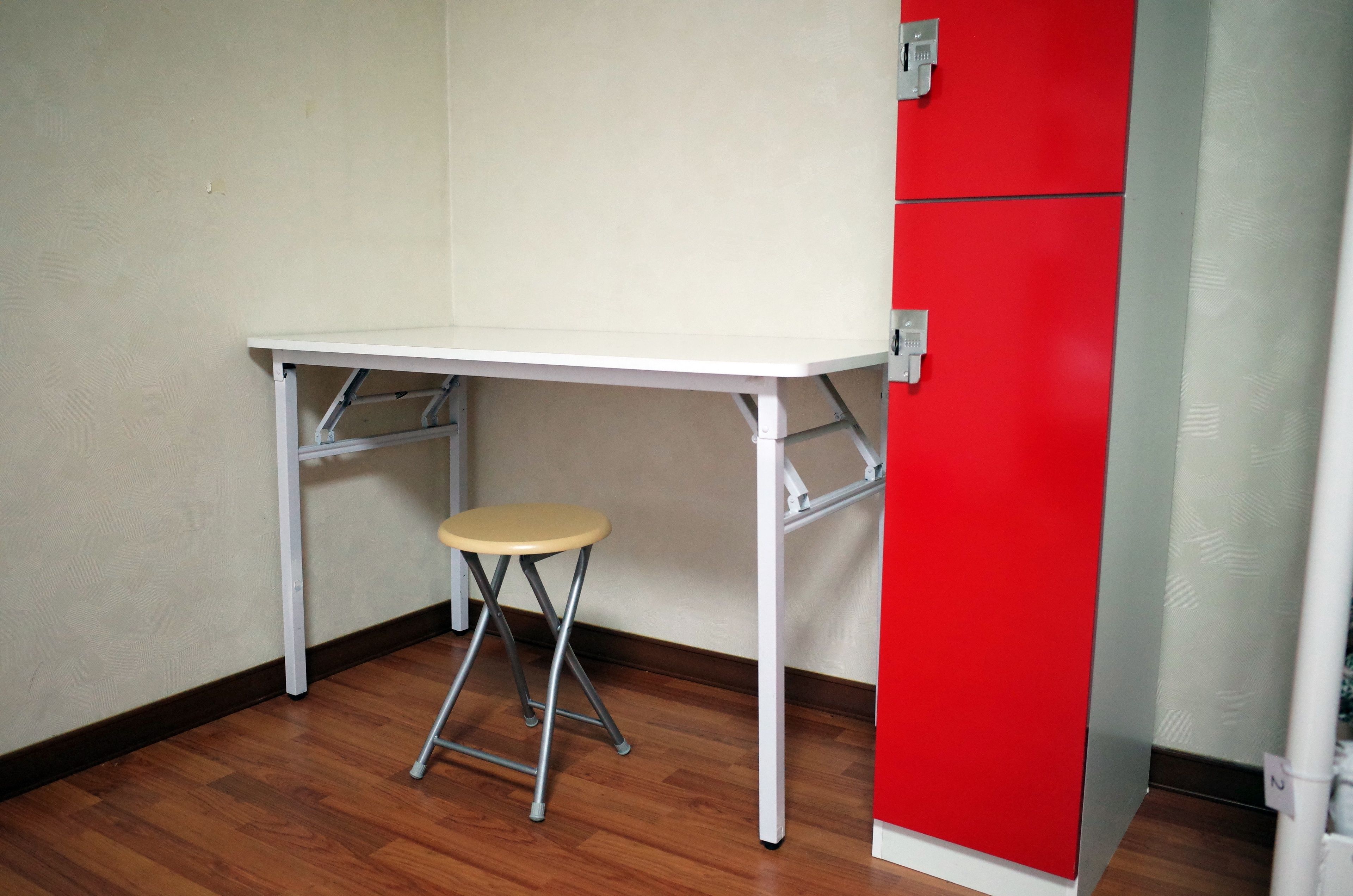 A corner room with a white desk and a stool, a red cabinet, and wooden flooring.