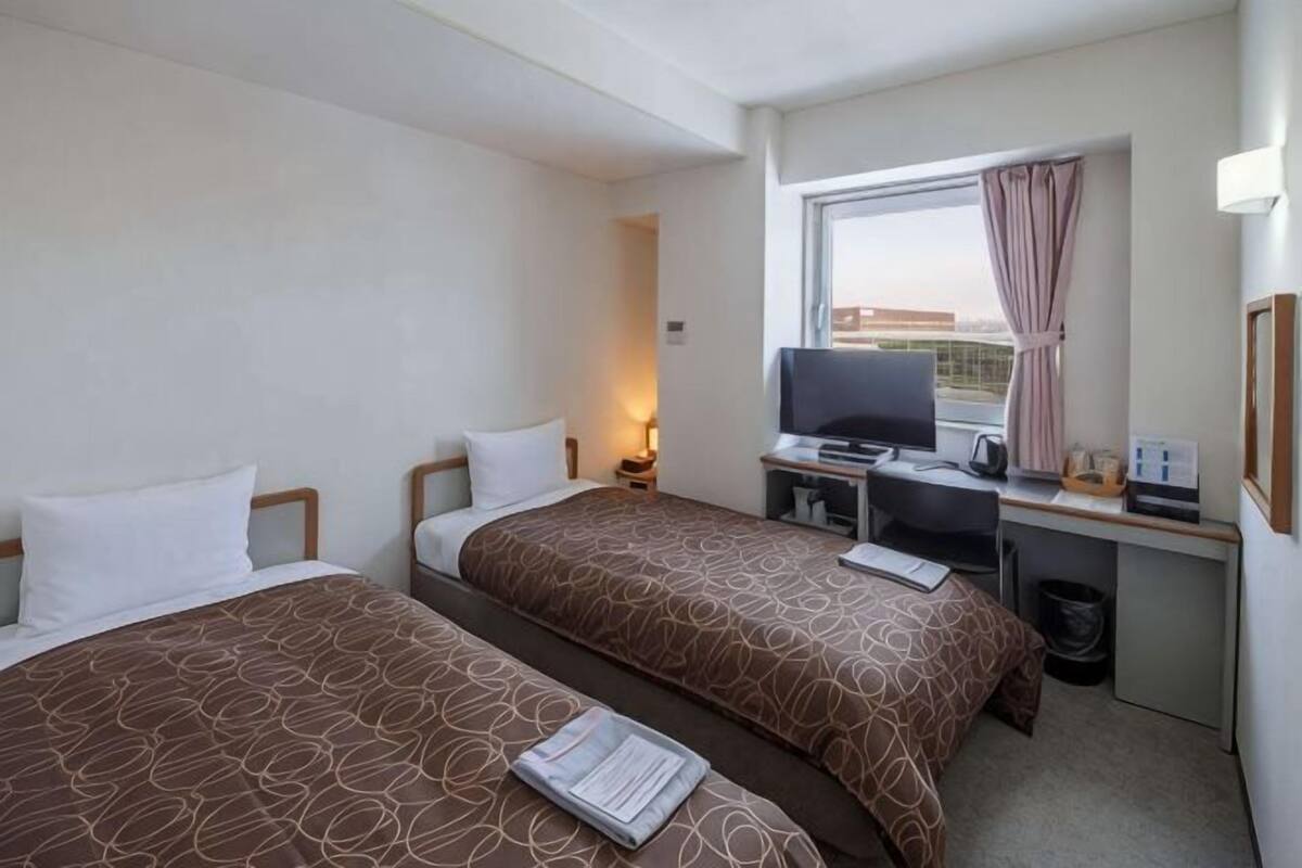 A hotel room with two beds, a desk with a computer, a TV, and a window with curtains.