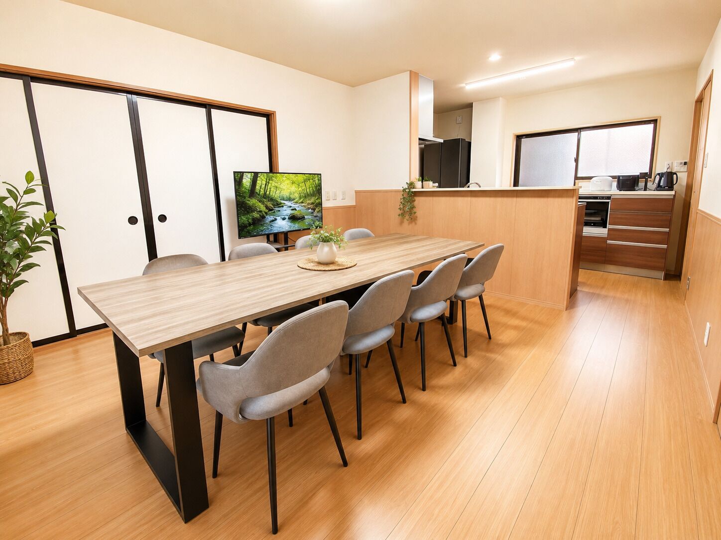 A modern dining area with a wooden table, grey chairs, a television, and a kitchen in the background.