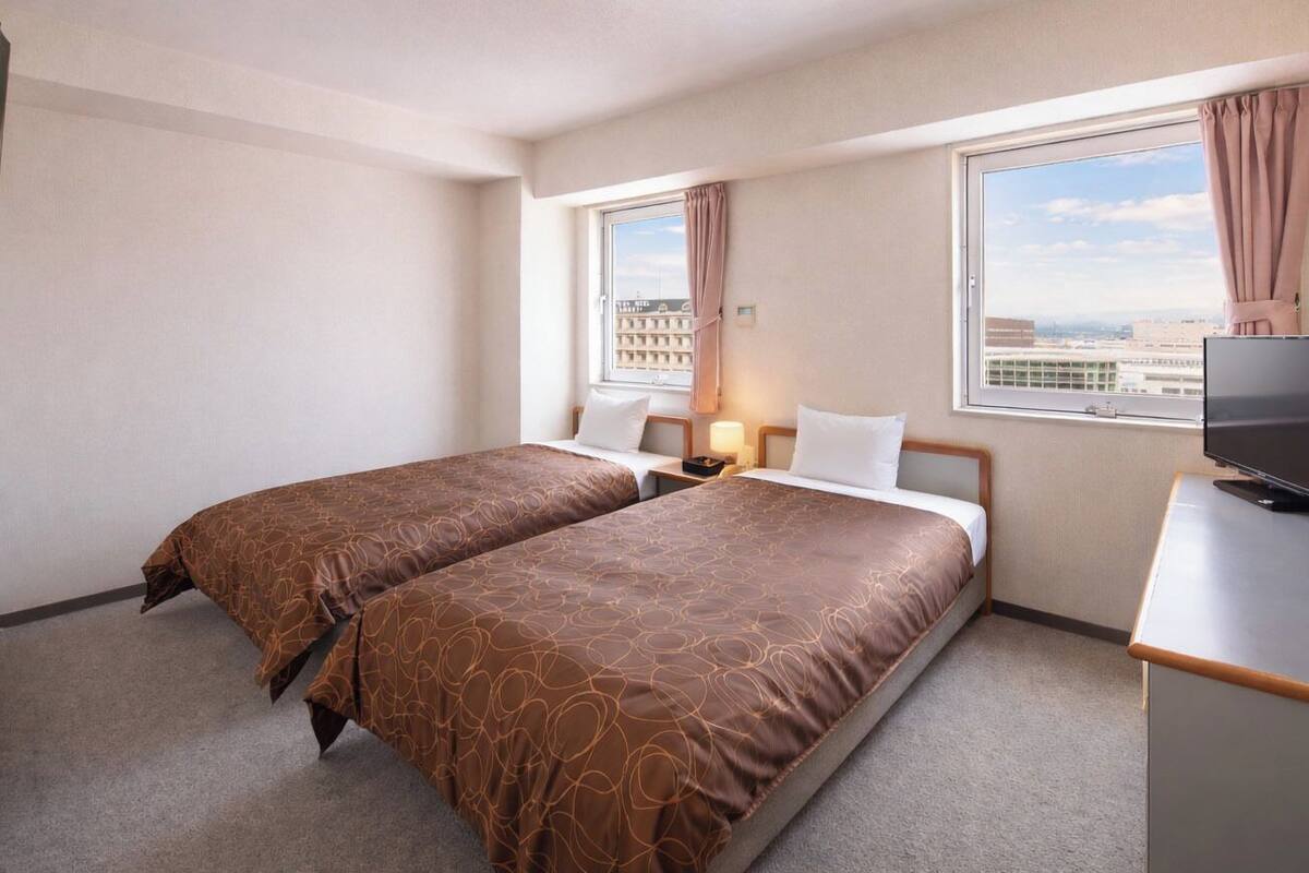 A hotel room with two beds, a TV, a desk, and a window with a city view.