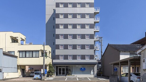 Un hotel de varios pisos con un diseño moderno, que cuenta con una entrada central con un letrero azul y un estacionamiento al frente.
