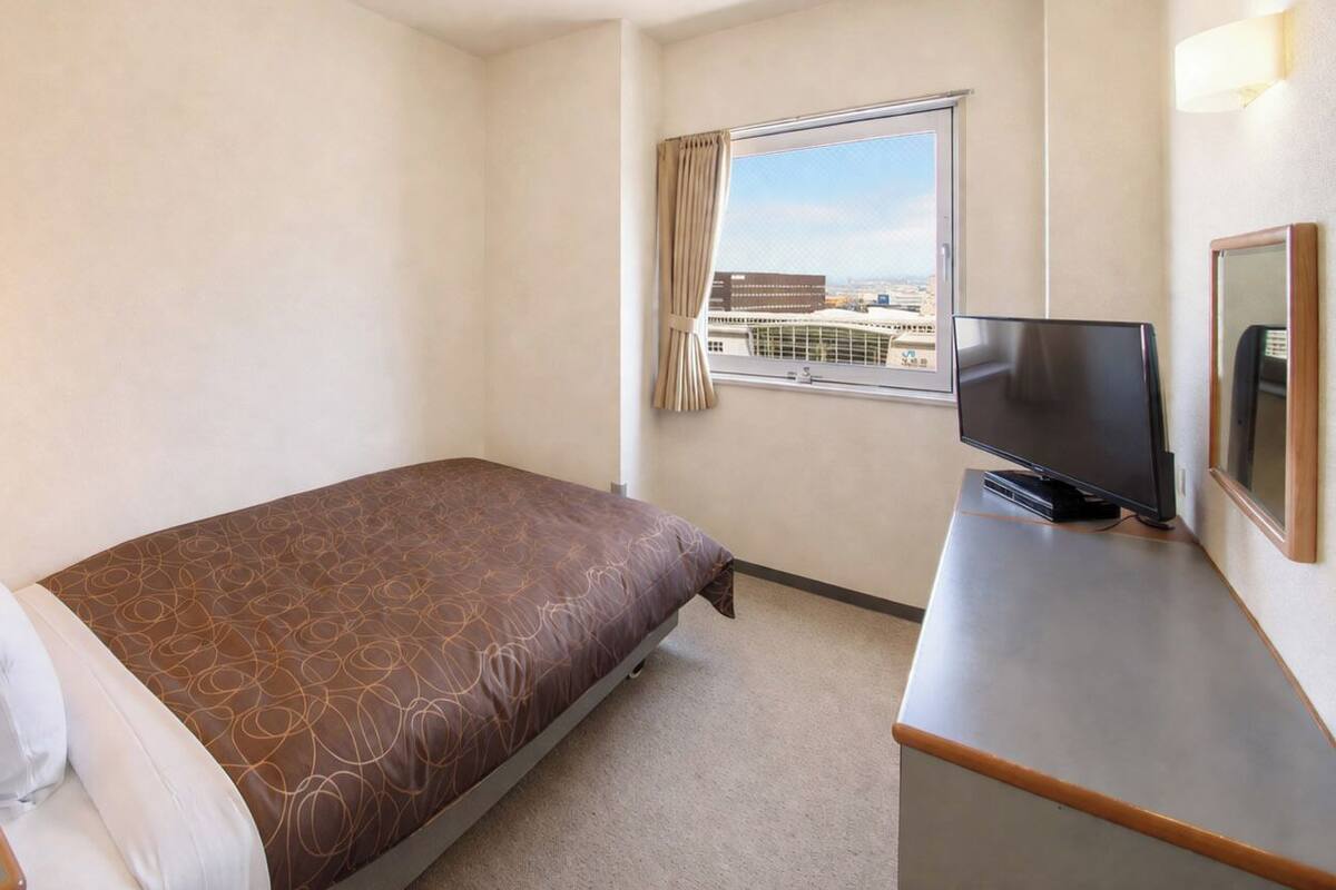 A hotel room with a bed, a desk with a television, a window with a city view, and a telephone.