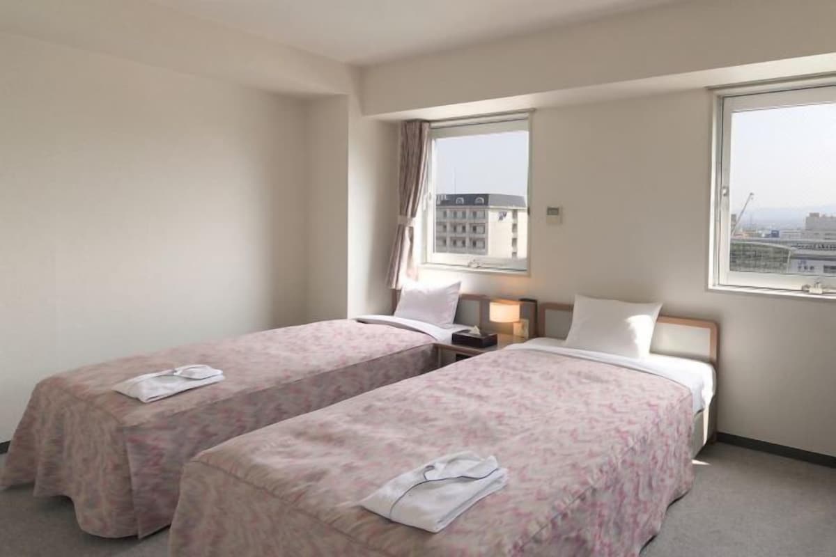 A hotel room with two beds, a nightstand, a window with curtains, and a view of buildings.