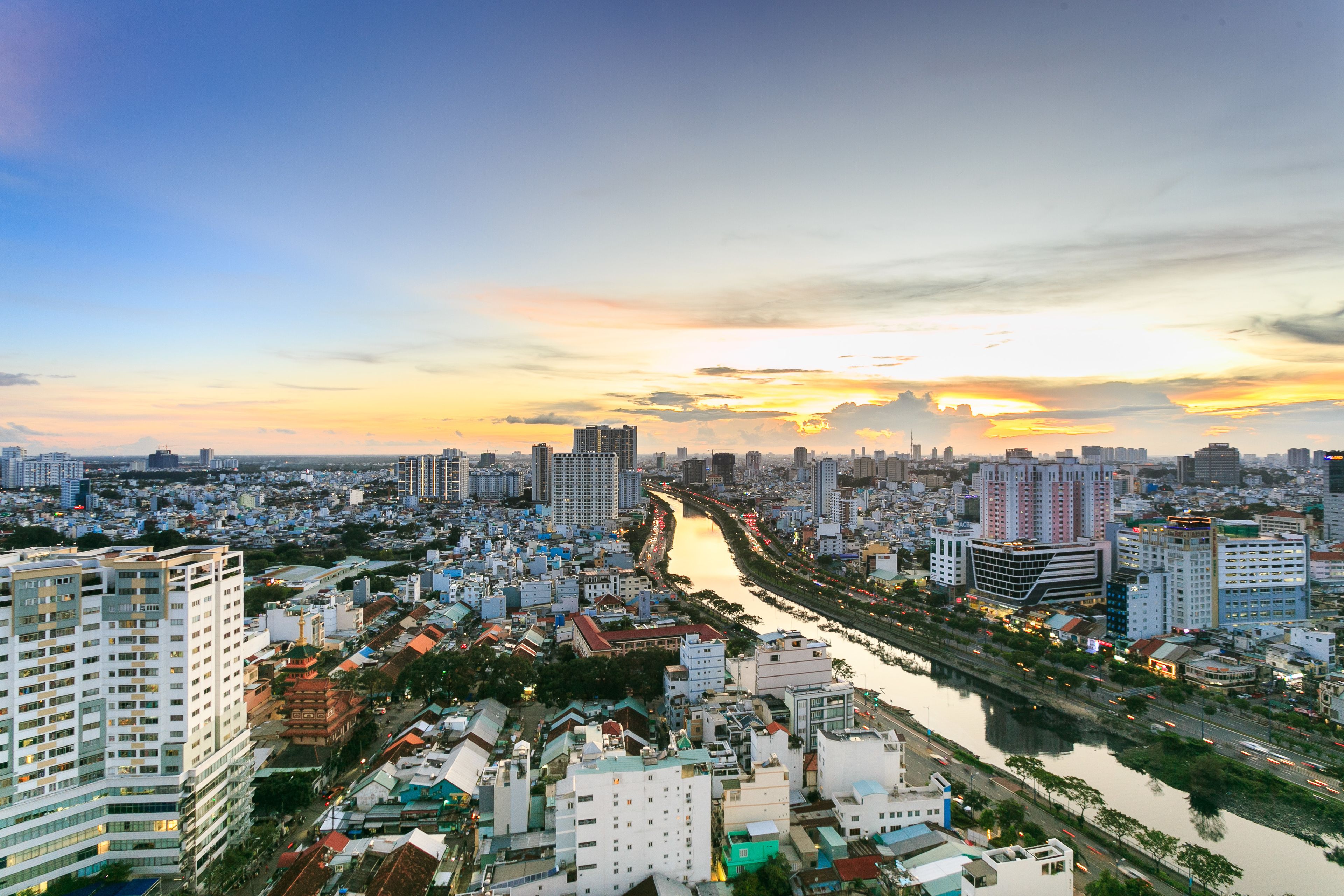 Une vue urbaine avec une rivière qui la traverse, entourée de différents bâtiments et un coucher de soleil en arrière-plan.