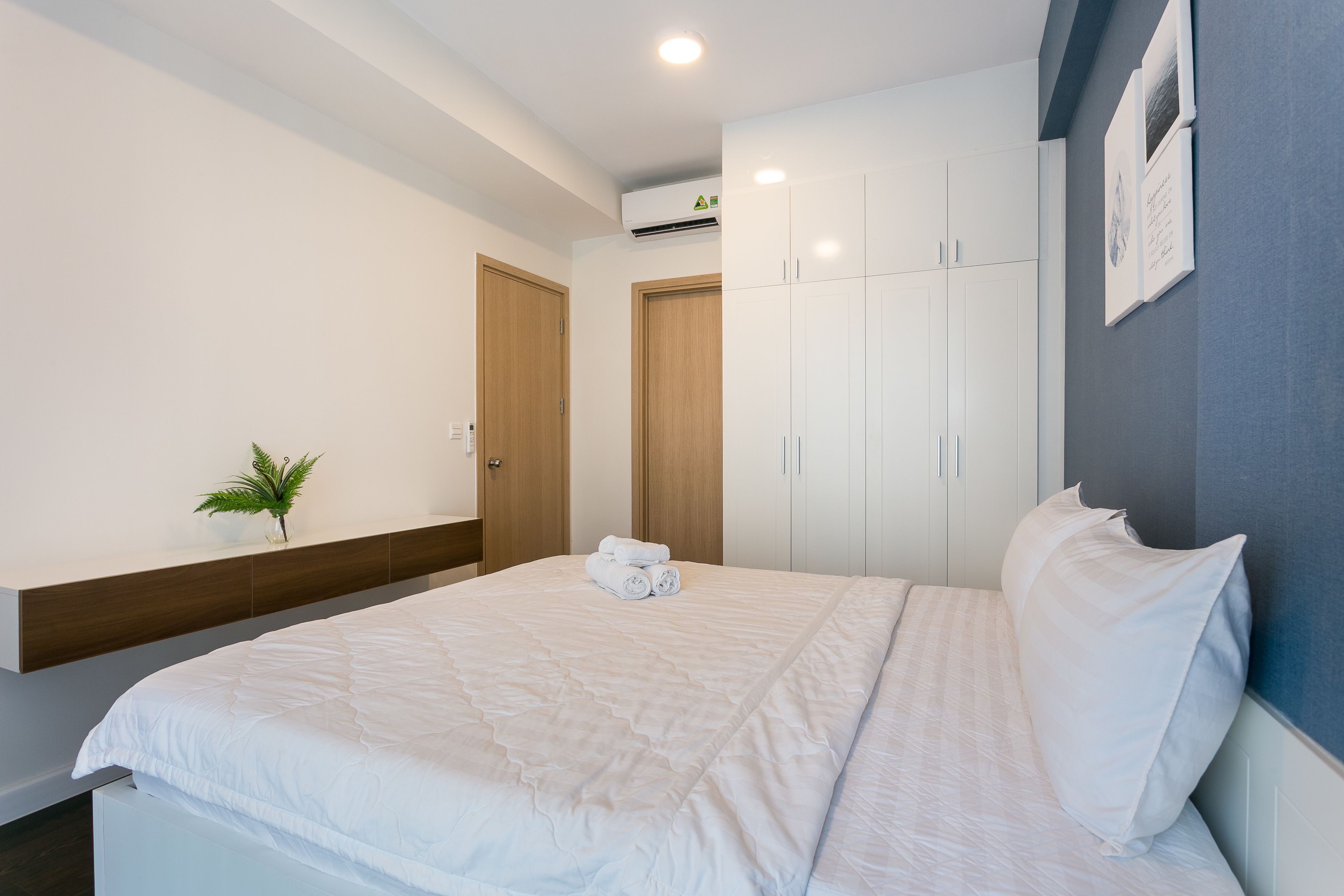 A neatly made bed with white linens, a small potted plant, and a set of white cabinets in a modern room with a grey accent wall.