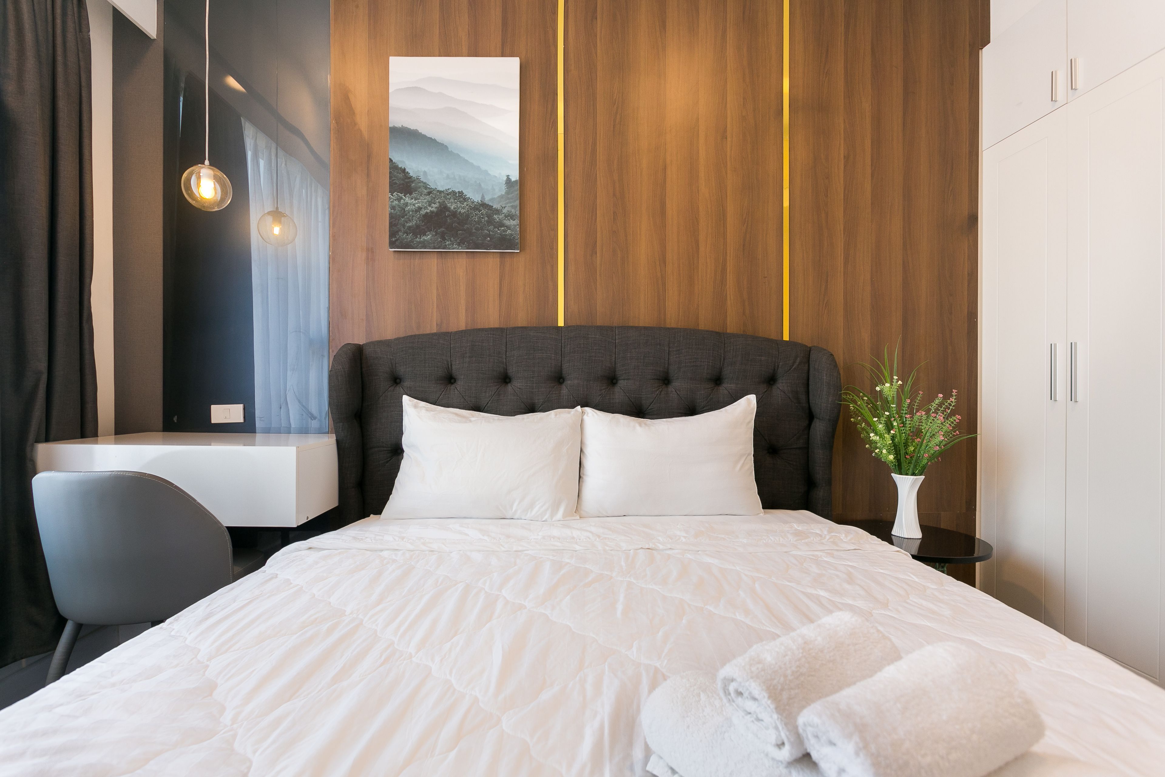 A neatly made bed with white bedding, a grey upholstered headboard, and a small bedside table with a vase of flowers.