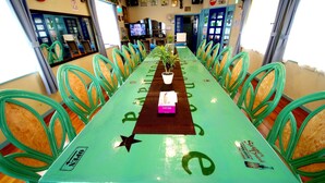 Uma mesa comprida com cadeiras em um quarto com uma televisão, um letreiro da Coca-Cola e uma placa de 'Sem Contato' na parede.