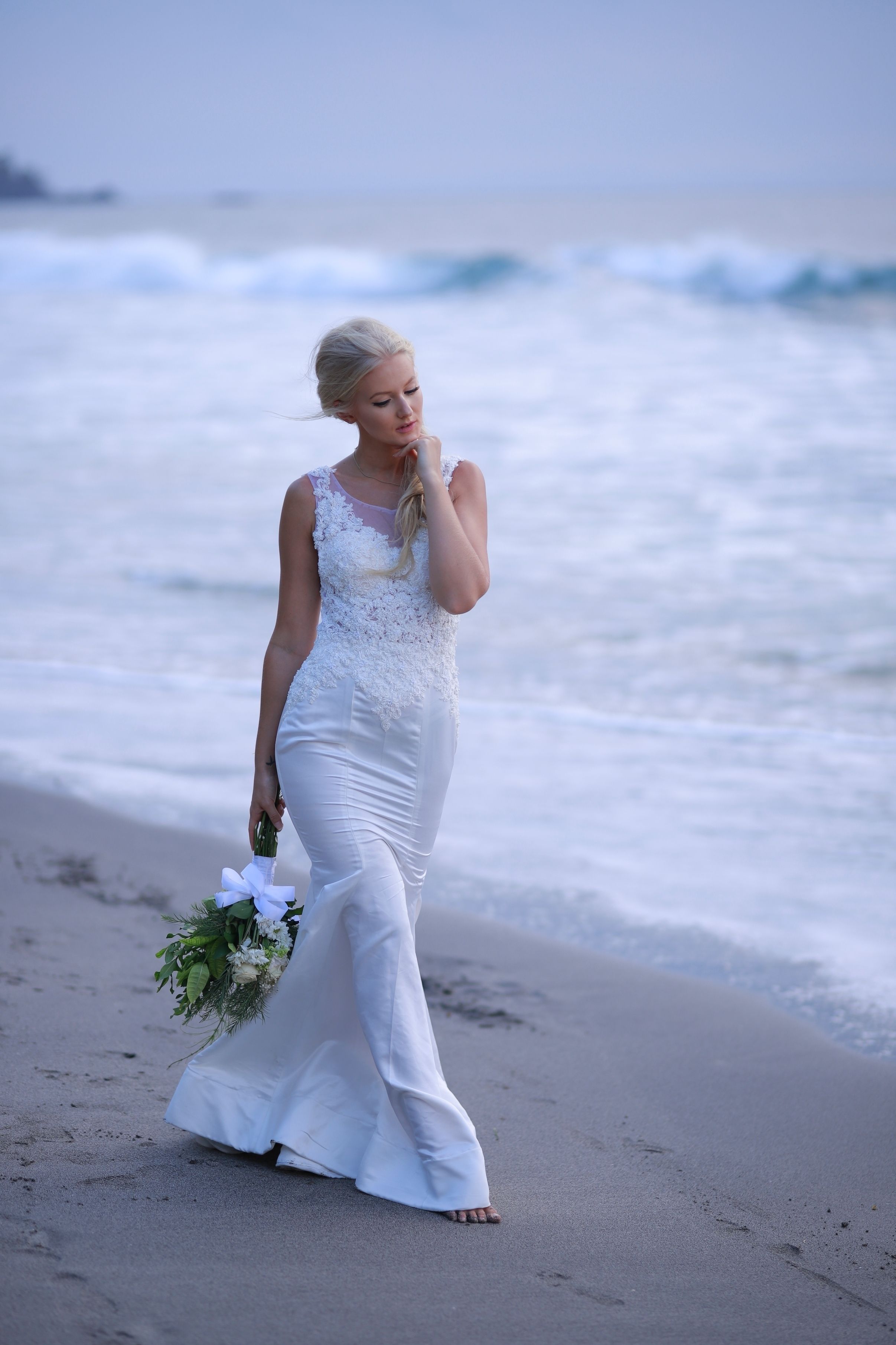 A bride in a white dress walking on a beach with ocean waves in the background.
