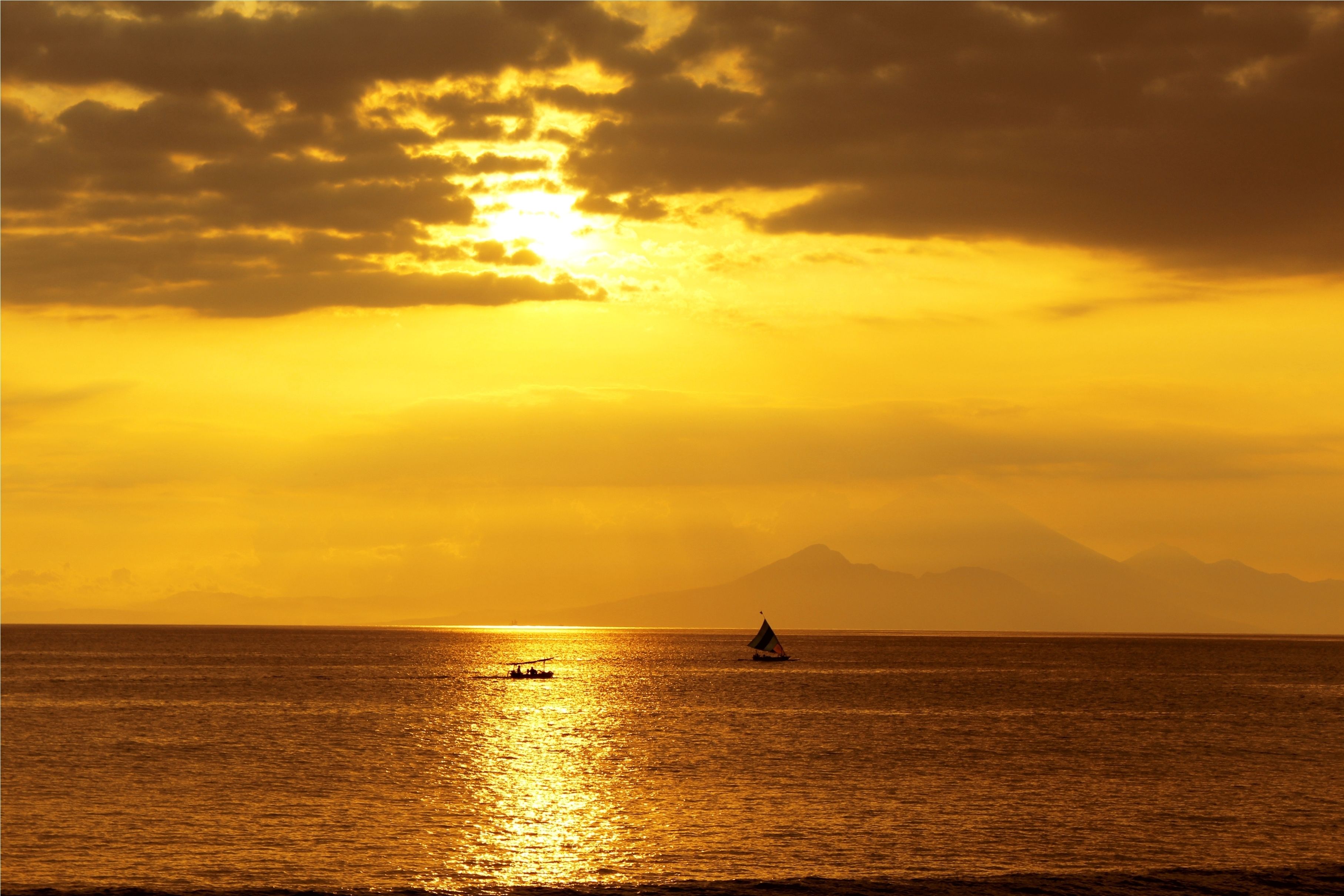 A sunset over a calm sea with a sailboat and a mountain in the distance.